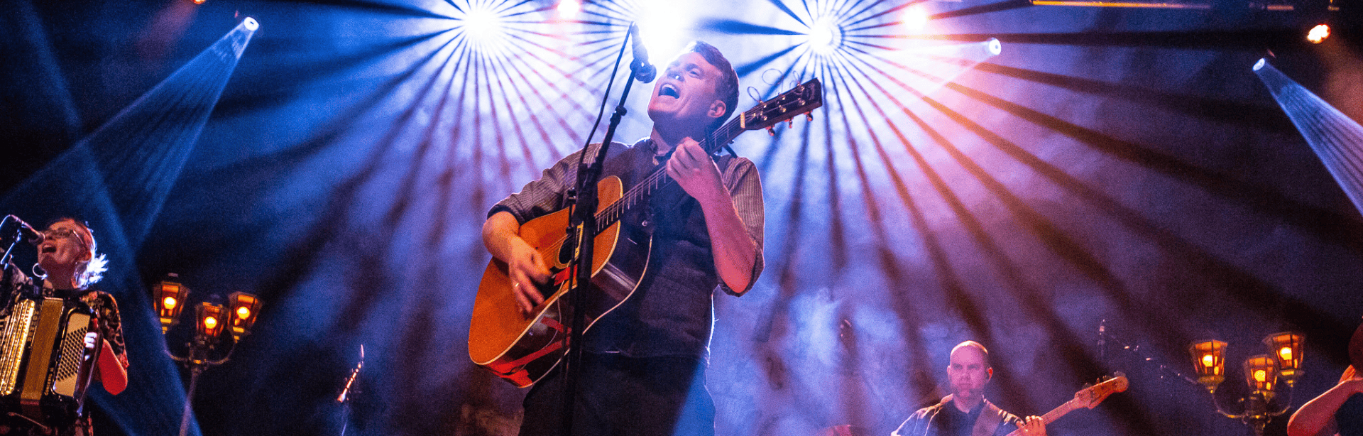 Mehrköpfige Band auf einer bunt beleuchteten Bühne, der Sänger spielt Gitarre und singt leidenschaftlich ins Mikrofon.